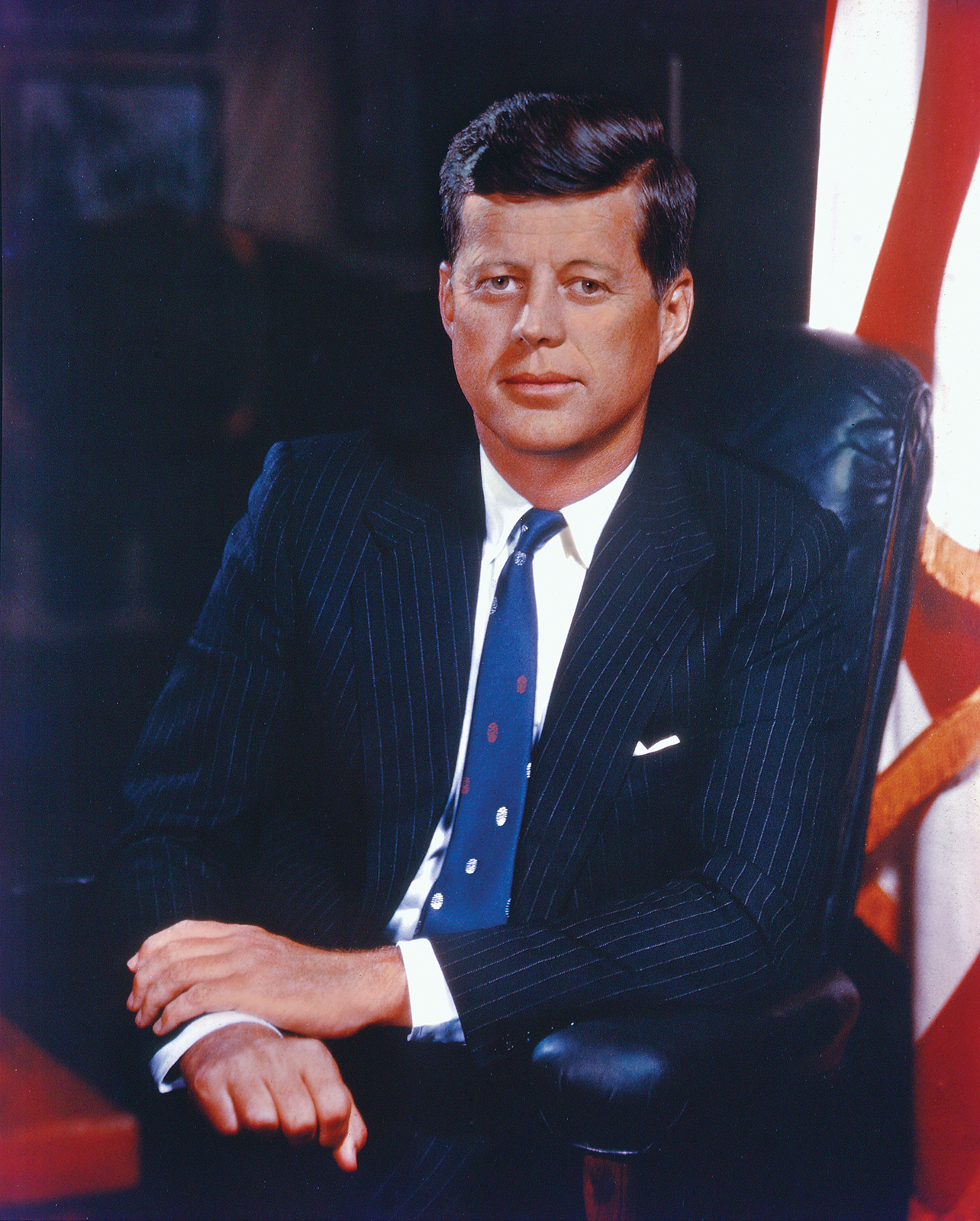 Presidential portrait of John F Kennedy dressed in a suit with a blue tie and next to the Flag of the United States.